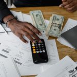Documentos financeiros, calculadora, dinheiro e laptop organizados sobre uma mesa de trabalho.
