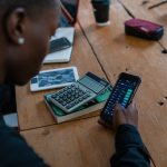 Pessoa usando um smartphone para analisar dados financeiros, com uma calculadora sobre a mesa.