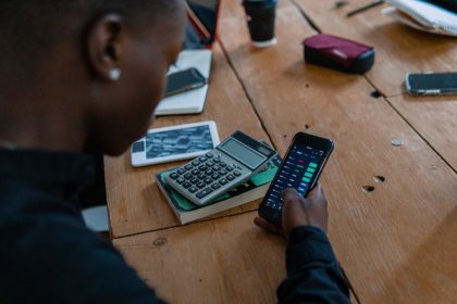 Pessoa usando um smartphone para analisar dados financeiros, com uma calculadora sobre a mesa.