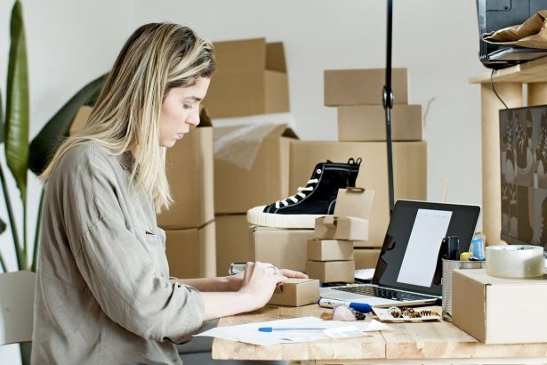 Microempreendedora embala caixas em um espaço de trabalho, cercada por materiais de envio e um laptop.