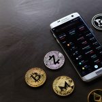 Moedas de criptomoeda ao lado de um smartphone que exibe uma plataforma digital de negociação.