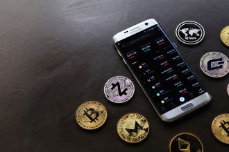 Moedas de criptomoeda ao lado de um smartphone que exibe uma plataforma digital de negociação.