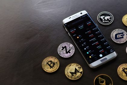 Moedas de criptomoeda ao lado de um smartphone que exibe uma plataforma digital de negociação.