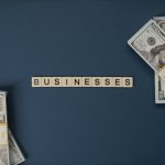 Cédulas de dólar dispostas ao lado de peças com a palavra “businesses”, em referência a negócios e transações financeiras.